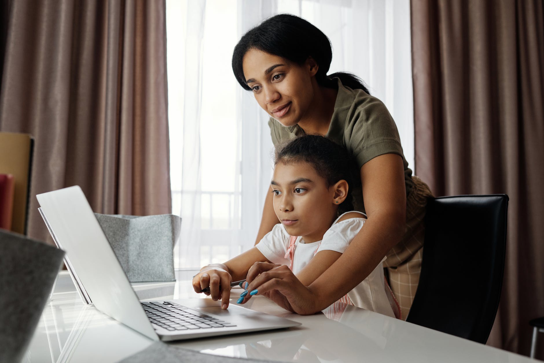 A child sitting at a table with laptop open, woman leaning over her helping to use the computer.