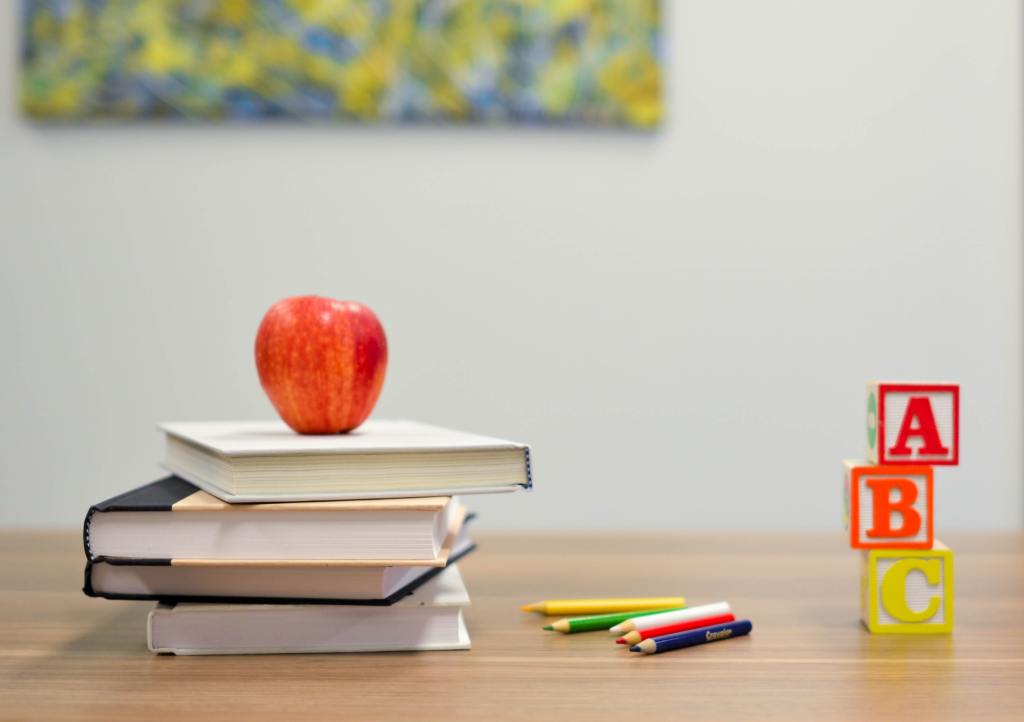 a stack of books with an apple on top, colored pencils and blocks with the letters ABC sitting on a desk