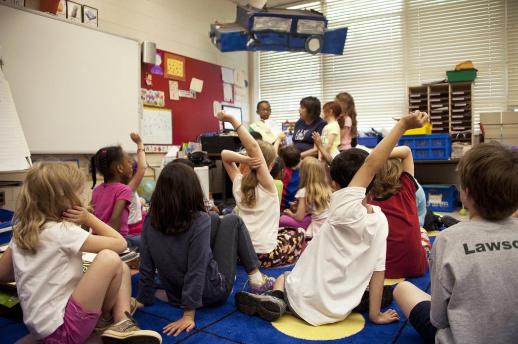 kids sitting on a rug. Some are raising their hand